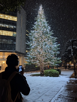 傍晚时分,日本的雪下得很大,而大雨刚刚停止。一棵高大的圣诞树在办公楼旁边亮着灯,闪耀着明亮的光芒。从后面拍照的人头上戴着耳机,穿着黑色夹克、白色衬衫和牛仔裤。他们拿出手机准备拍摄。混凝土路的两边还留有冬天的泥渍,在大树的附近可以看到一些小水坑。这张照片是在夜晚使用广角镜头拍摄的,采用了日本艺术家的风格。