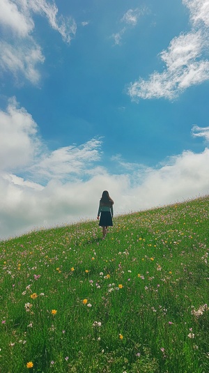 一半画面一个女人走在开满鲜花的绿色草原，画面一半天空蓝天白云