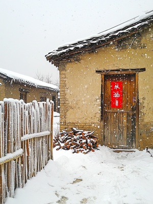 在冬季,天空多云且沉重地覆盖着雪。在中国农村的一座土坯房前,一个木栅栏矗立在白色的、被雪覆盖的泥墙之间。门上的红色对联为外观增添了色彩。下面还有一些柴火,为人们在穿越雪景时的行走增添了温暖。这张照片是用手机相机拍摄的。它有一个广角镜头,突出了每栋建筑的每一个细节。感觉就像你可以感受到皮肤上的冷空气,甚至在你的...