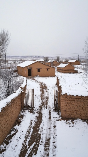 冬季中央的一座小土坯房,村庄，几处房子，屋顶和墙壁覆盖着雪,房子正前面有泥墙，栅栏大门，一座标准的农家小院，泥土路、覆盖着雪的地面和灰暗的天空,使用iPhone相机拍摄。该场景在无人机镜头下拍摄，鸟瞰图